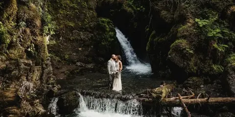 A bride and groom in wedding attire standing together in front of a forest waterfall.