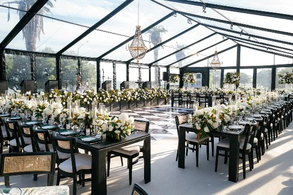 A luxury wedding reception venue featuring a clear tent, long tables with white floral arrangements, and hanging chandeliers.