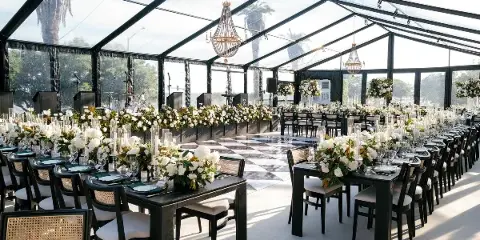 A luxury wedding reception venue featuring a clear tent, long tables with white floral arrangements, and hanging chandeliers.