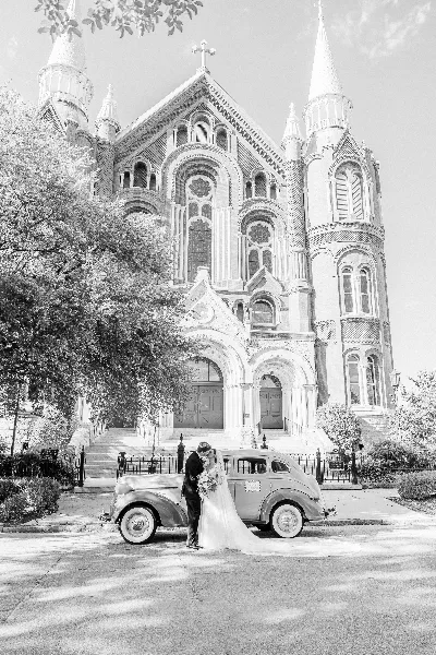 Black-and-white wedding photo of a bride and groom kissing beside a vintage car in front of a church.