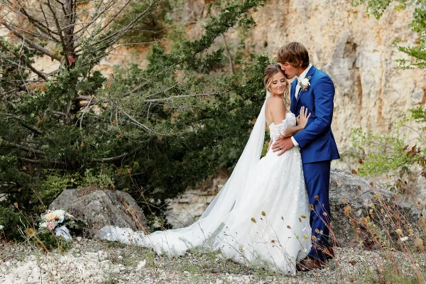 A bride in a white lace gown and a groom in a blue suit embracing outdoors near a rocky cliff face.