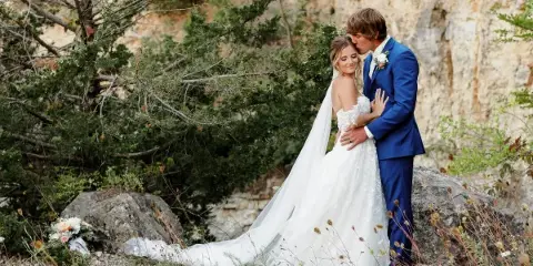 A bride in a white lace gown and a groom in a blue suit embracing outdoors near a rocky cliff face.
