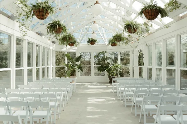 Interior of a white Victorian greenhouse wedding venue with rows of white chairs, hanging ferns, and a glass ceiling.