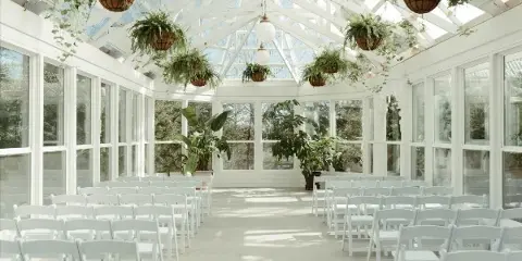 Interior of a white Victorian greenhouse wedding venue with rows of white chairs, hanging ferns, and a glass ceiling.