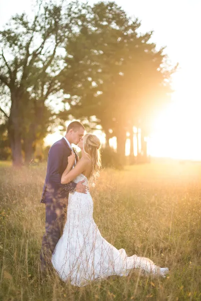 A bride and groom embracing in a grassy field during golden hour with trees in the background.