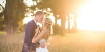A bride and groom embracing in a grassy field during golden hour with trees in the background.