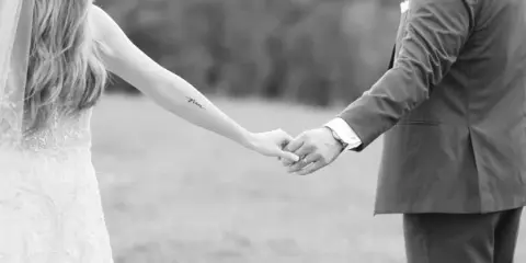 A black and white photograph of a bride and groom holding hands in an outdoor setting.