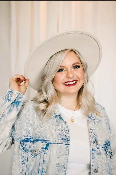 A smiling woman with styled blonde hair and professional makeup, wearing a denim jacket and a wide-brimmed hat.