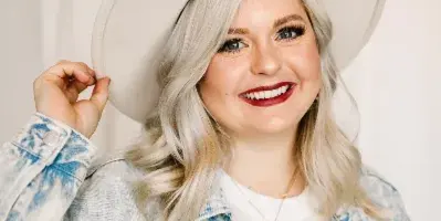 A smiling woman with styled blonde hair and professional makeup, wearing a denim jacket and a wide-brimmed hat.