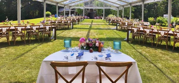 Outdoor wedding reception setup featuring a long table with floral arrangements and chairs under a clear tent.