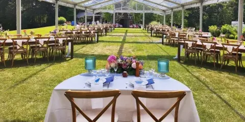 Outdoor wedding reception setup featuring a long table with floral arrangements and chairs under a clear tent.