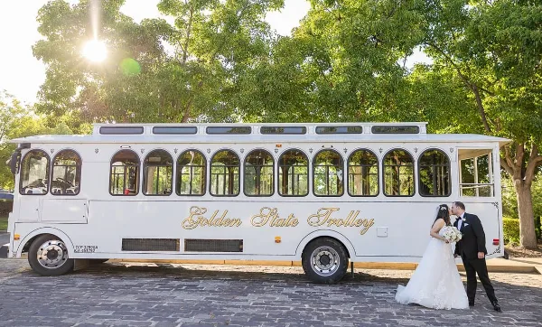 A white luxury trolley bus with 'Golden State Trolley' written on the side, parked outdoors with a bride and groom standing in front of it.