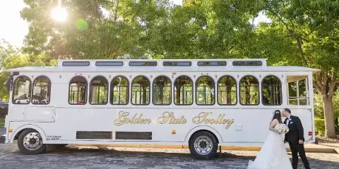 A white luxury trolley bus with 'Golden State Trolley' written on the side, parked outdoors with a bride and groom standing in front of it.
