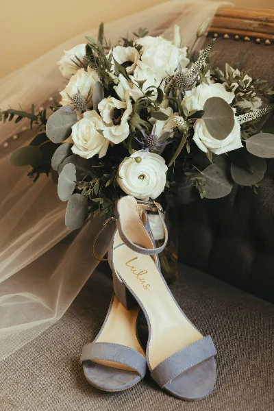 A close-up of a white bridal bouquet with eucalyptus and ranunculus resting on a chair next to a pair of grey high-heeled shoes.