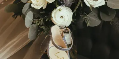 A close-up of a white bridal bouquet with eucalyptus and ranunculus resting on a chair next to a pair of grey high-heeled shoes.