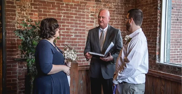 A wedding officiant in a grey suit stands between a bride and groom, holding a book during a ceremony in a brick-walled venue.