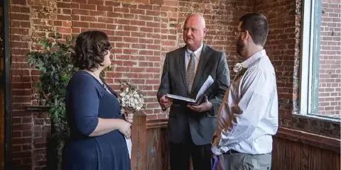 A wedding officiant in a grey suit stands between a bride and groom, holding a book during a ceremony in a brick-walled venue.