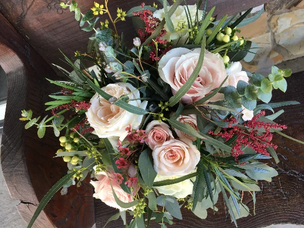 A close-up of a romantic floral arrangement with blush and white roses, eucalyptus, and red accents resting on a wooden surface.