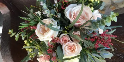 A close-up of a romantic floral arrangement with blush and white roses, eucalyptus, and red accents resting on a wooden surface.