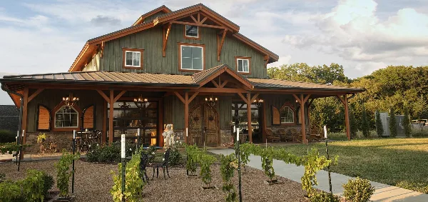 Exterior view of a rustic green barn-style event venue with a stone base, wooden beams, and young grapevines planted in the foreground.