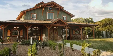 Exterior view of a rustic green barn-style event venue with a stone base, wooden beams, and young grapevines planted in the foreground.