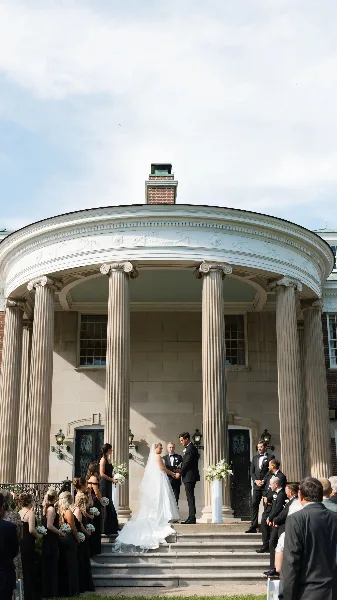 A bride and groom exchanging vows on the steps of a classical building with columns, surrounded by their wedding party.
