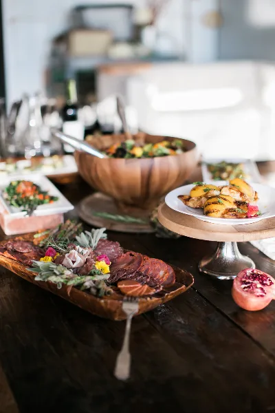 A catering table with a wooden salad bowl, charcuterie board, plated appetizers, and a pomegranate on a rustic wooden surface.