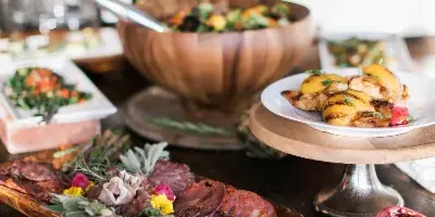 A catering table with a wooden salad bowl, charcuterie board, plated appetizers, and a pomegranate on a rustic wooden surface.
