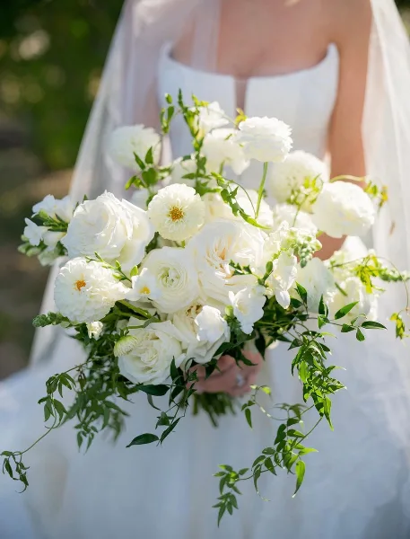A close-up of a bride holding a large white floral bouquet featuring roses, ranunculus, and greenery.