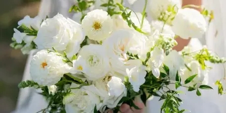 A close-up of a bride holding a large white floral bouquet featuring roses, ranunculus, and greenery.