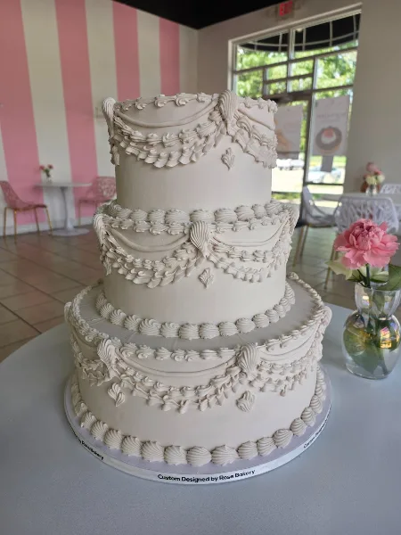 A three-tier white wedding cake with intricate piped floral decorations, displayed in a pink and white striped room with a vase of pink flowers.