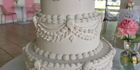 A three-tier white wedding cake with intricate piped floral decorations, displayed in a pink and white striped room with a vase of pink flowers.