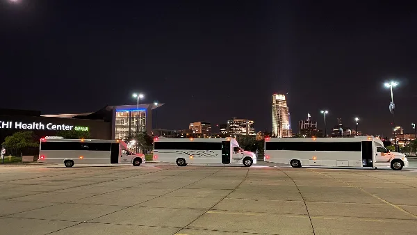 Three white shuttle buses parked in a lot at night with the Omaha skyline and CHI Health Center in the background.