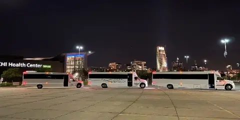 Three white shuttle buses parked in a lot at night with the Omaha skyline and CHI Health Center in the background.
