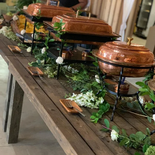 Catering buffet table with copper chafing dishes, white flowers, and serving utensils on a wooden table.