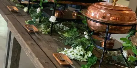 Catering buffet table with copper chafing dishes, white flowers, and serving utensils on a wooden table.