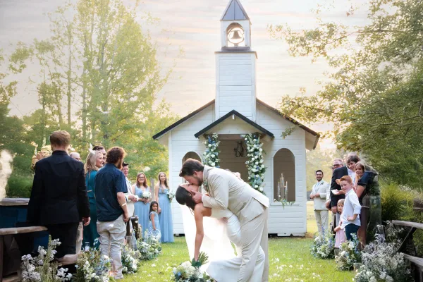 A bride and groom kissing during a wedding ceremony in front of a white chapel with a bell tower.