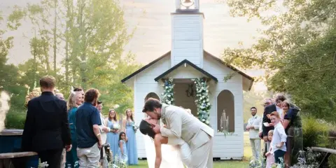 A bride and groom kissing during a wedding ceremony in front of a white chapel with a bell tower.