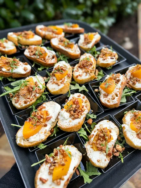A black serving tray filled with crostini appetizers topped with white cheese, orange fruit, and nuts.