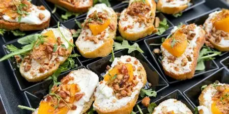 A black serving tray filled with crostini appetizers topped with white cheese, orange fruit, and nuts.