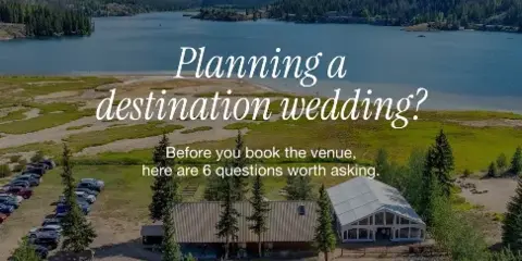 Aerial view of a rustic wedding venue building situated on the shore of a large lake with mountains in the background.