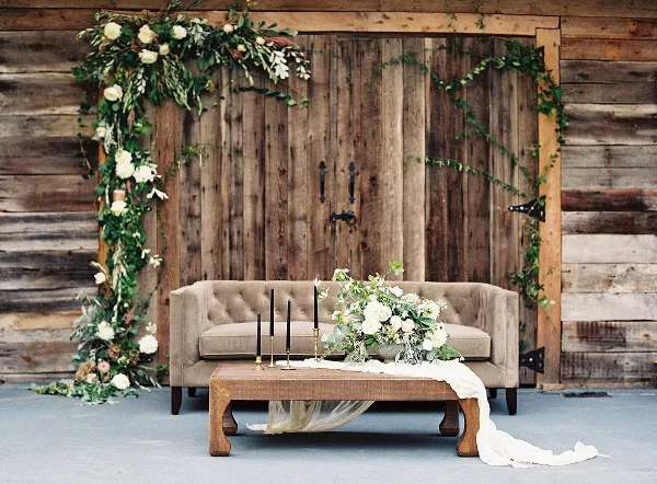A rustic wooden backdrop adorned with white floral garlands, featuring a beige sofa and a wooden coffee table with candles and tulle.