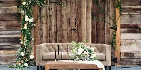 A rustic wooden backdrop adorned with white floral garlands, featuring a beige sofa and a wooden coffee table with candles and tulle.