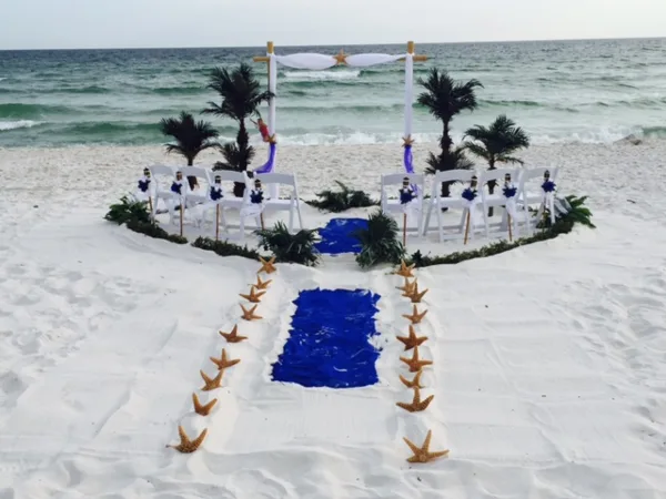 Beach wedding ceremony setup with white chairs, sashes, starfish-lined aisle, and a draped arch near the ocean