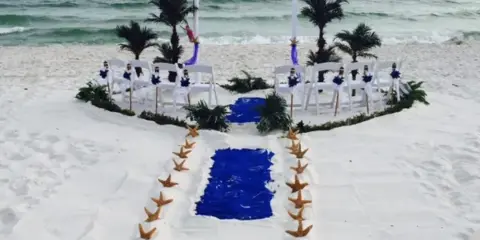 Beach wedding ceremony setup with white chairs, sashes, starfish-lined aisle, and a draped arch near the ocean