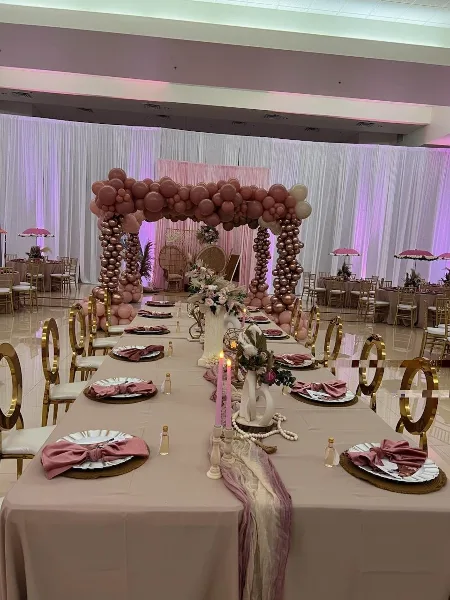 A long event table set with beige linens, gold chairs, plates, napkins, candles, and floral centerpieces in front of a pink and gold balloon arch.
