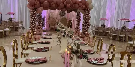 A long event table set with beige linens, gold chairs, plates, napkins, candles, and floral centerpieces in front of a pink and gold balloon arch.