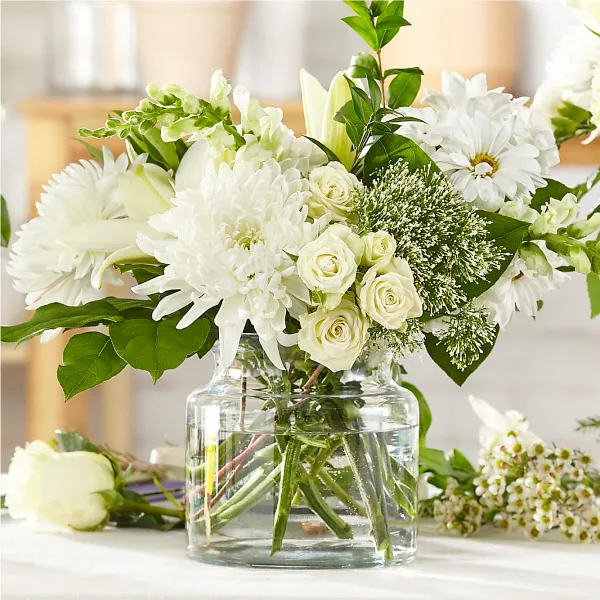 A white floral arrangement with roses, chrysanthemums, and lilies in a clear glass vase.