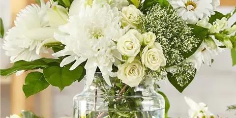 A white floral arrangement with roses, chrysanthemums, and lilies in a clear glass vase.