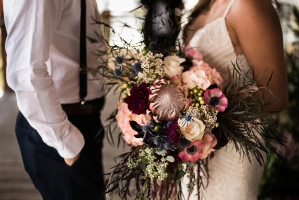 A bride in a white lace dress holding a large, lush wedding bouquet with pink and white flowers and dark greenery.